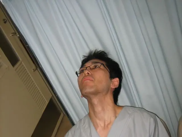 There is a person sitting on a chair and wearing specs. Behind him there is a curtain. Near to him...